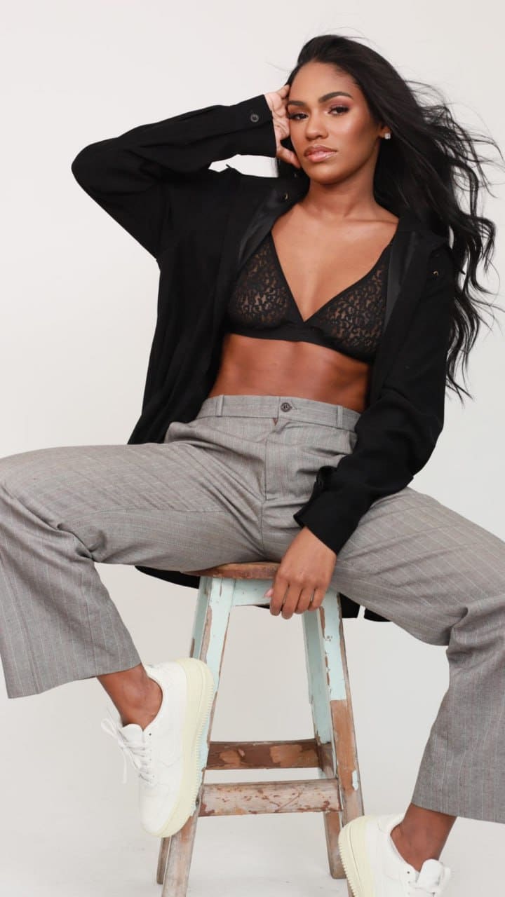 Sitting on a wooden stool against white, a woman wears an open black shirt, black lace bra, pinstripe trousers, and sneakers.