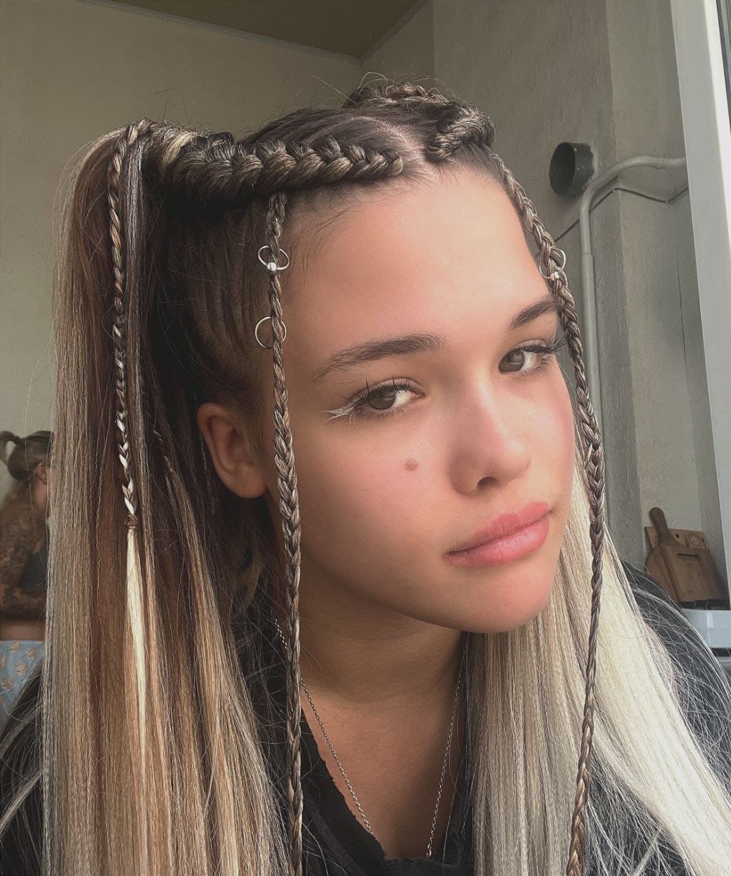 Jenia, a young woman with highlighted braids and a ponytail, wears a dark top and necklace near an indoor window.
