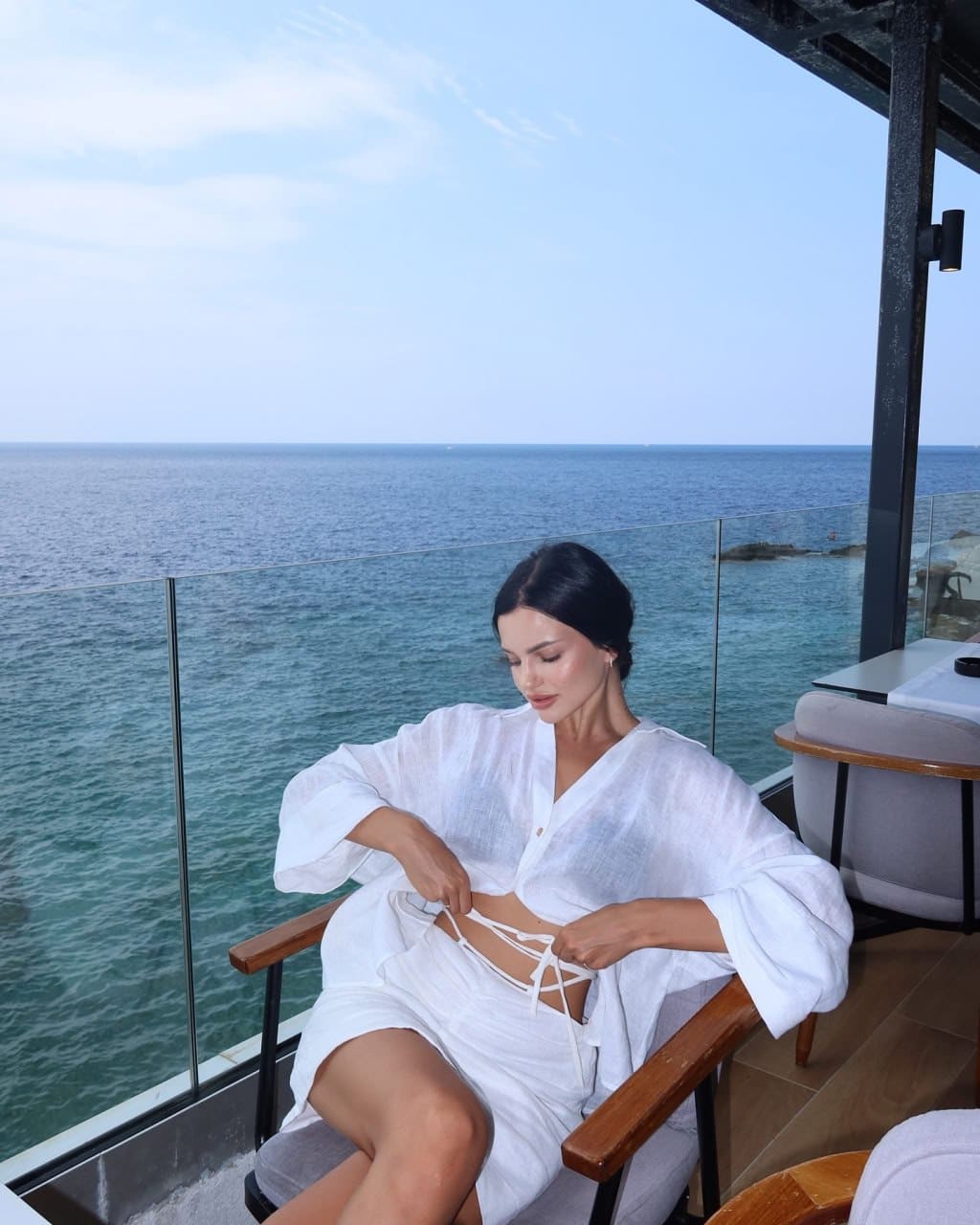Woman seated in a wooden chair on a glass-railed terrace, adjusting the lacing of an open white linen shirt over a strappy top, sea view behind.