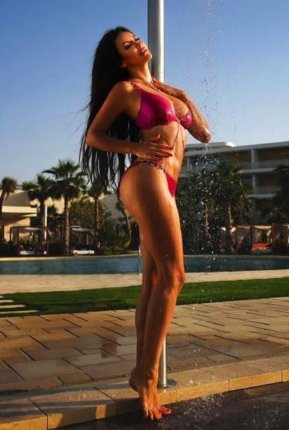 Barefoot on a tiled poolside deck, a woman in a pink bikini tilts her head back under water spray, with palm trees and a resort building behind her.