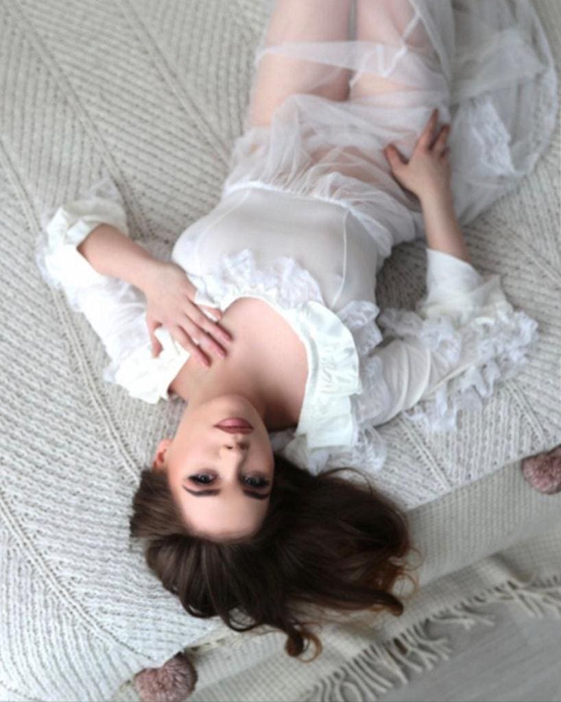 Lying on her back on a textured white blanket, she wears a sheer white ruffled top and skirt, one hand on her chest, one on her thigh.