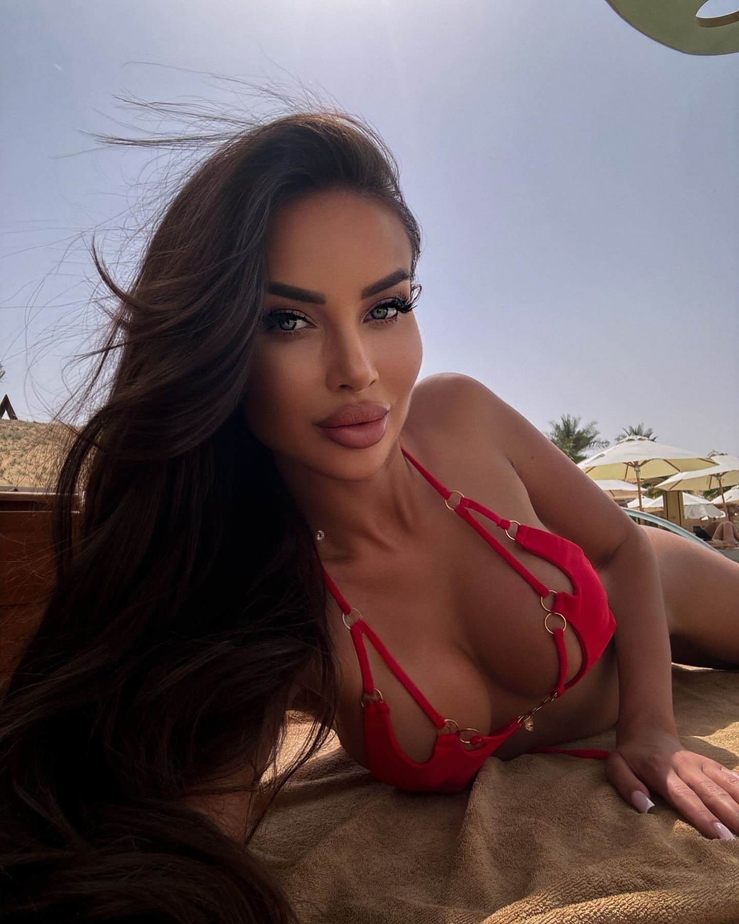 A woman lies on a tan towel outdoors in a red strappy bikini, with beach umbrellas and clear sky behind her.