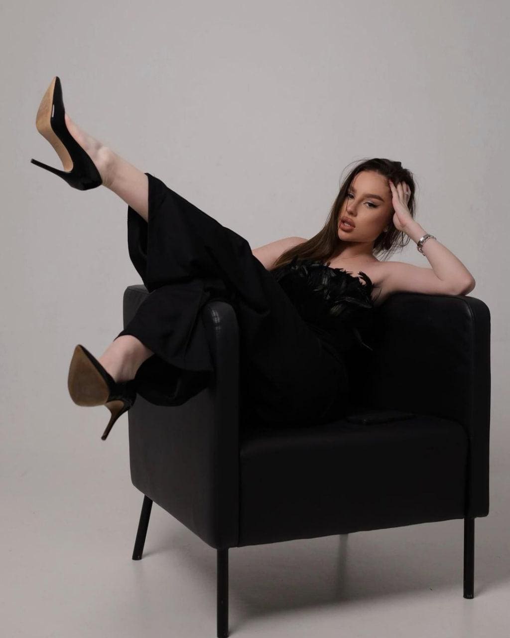 A woman in a strapless feathered top and wide-leg trousers reclines in a black armchair against a gray studio backdrop.