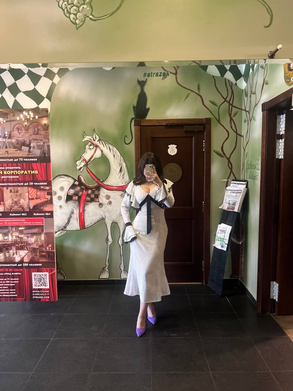 In a hallway with a horse mural, a woman in a white patterned midi dress and purple heels takes a mirror selfie.