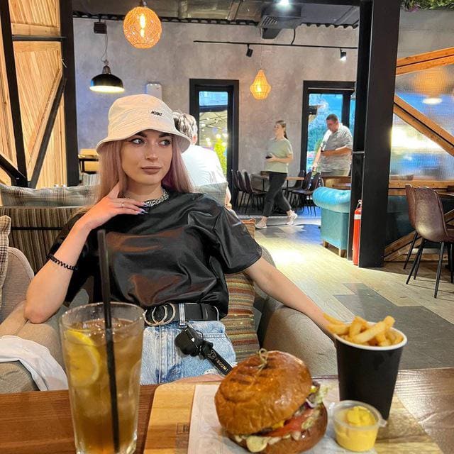 Alice sits at a restaurant table in a bucket hat, black top, and denim shorts, with a burger and fries in front of her.
