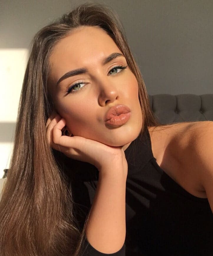 Resting her chin on her hand with a puckered-lip expression, she wears a black sleeveless top in front of a gray tufted headboard.