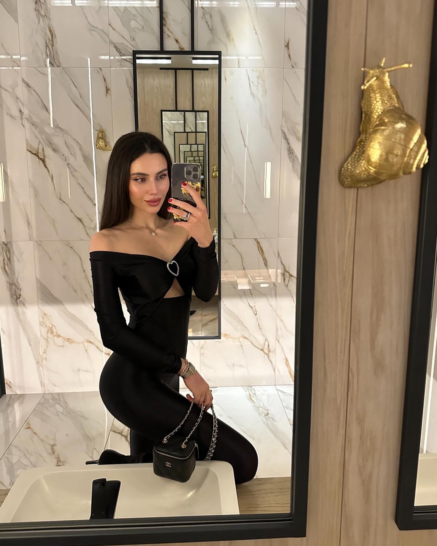 A woman takes a mirror selfie in a marble bathroom wearing a black off-the-shoulder bodysuit with a waist cutout, holding a black chain bag.