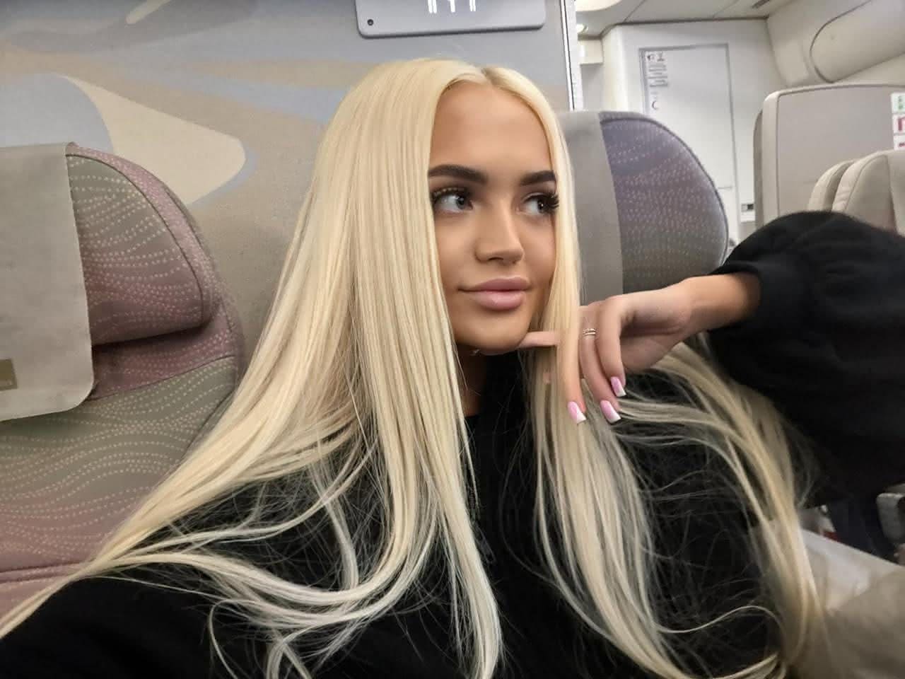 A woman in a black long-sleeved top sits in an airplane seat, resting her chin on her hand and looking aside.