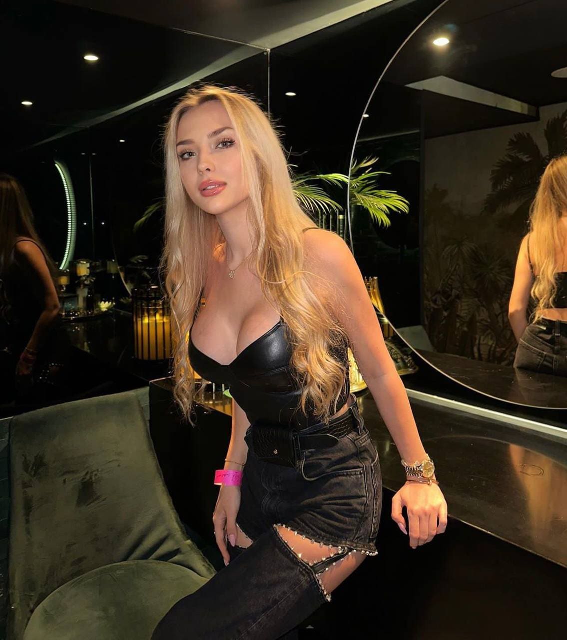 A woman with wavy blonde hair sits in a dimly lit room with mirrors and plants, wearing a black leather bustier, ripped denim shorts, and a pink wristband.
