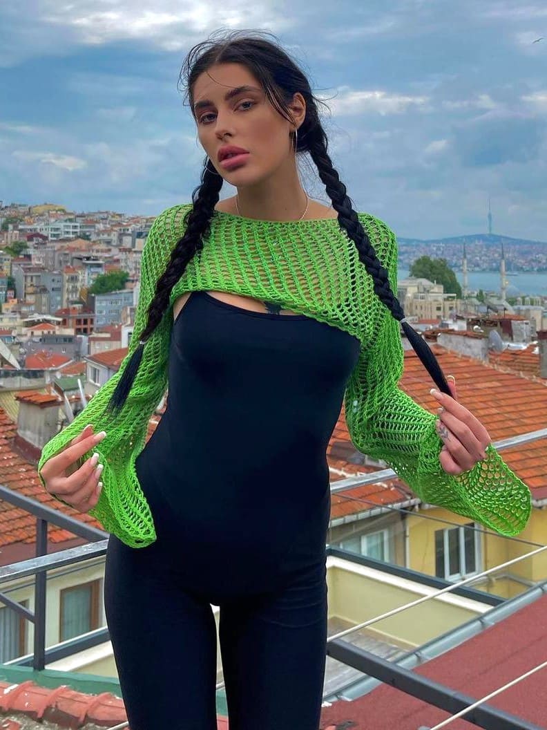 On a rooftop terrace, she wears a black bodysuit under a green open-knit cardigan, with a cityscape behind.
