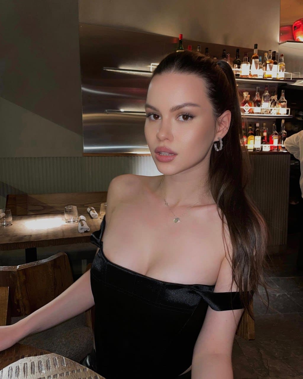 A woman with a high ponytail sits at a bar table in a black off-the-shoulder top and thin necklace, with shelves of liquor bottles behind her.