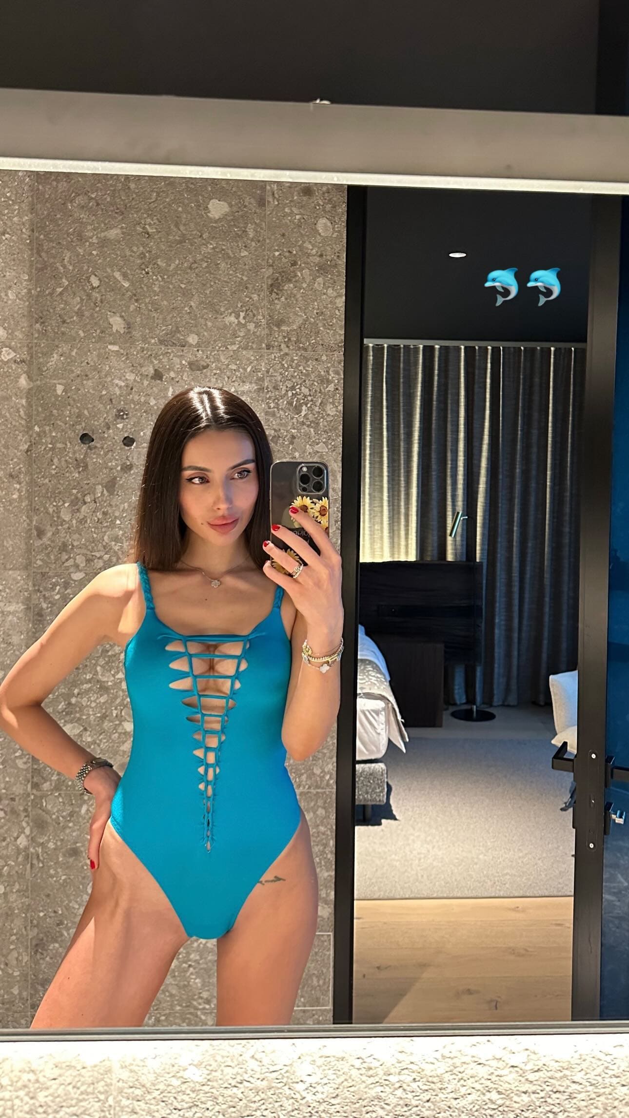 A mirror selfie captures a woman in a teal one-piece swimsuit with a lace-up front, standing in a room with a stone wall and bed visible.