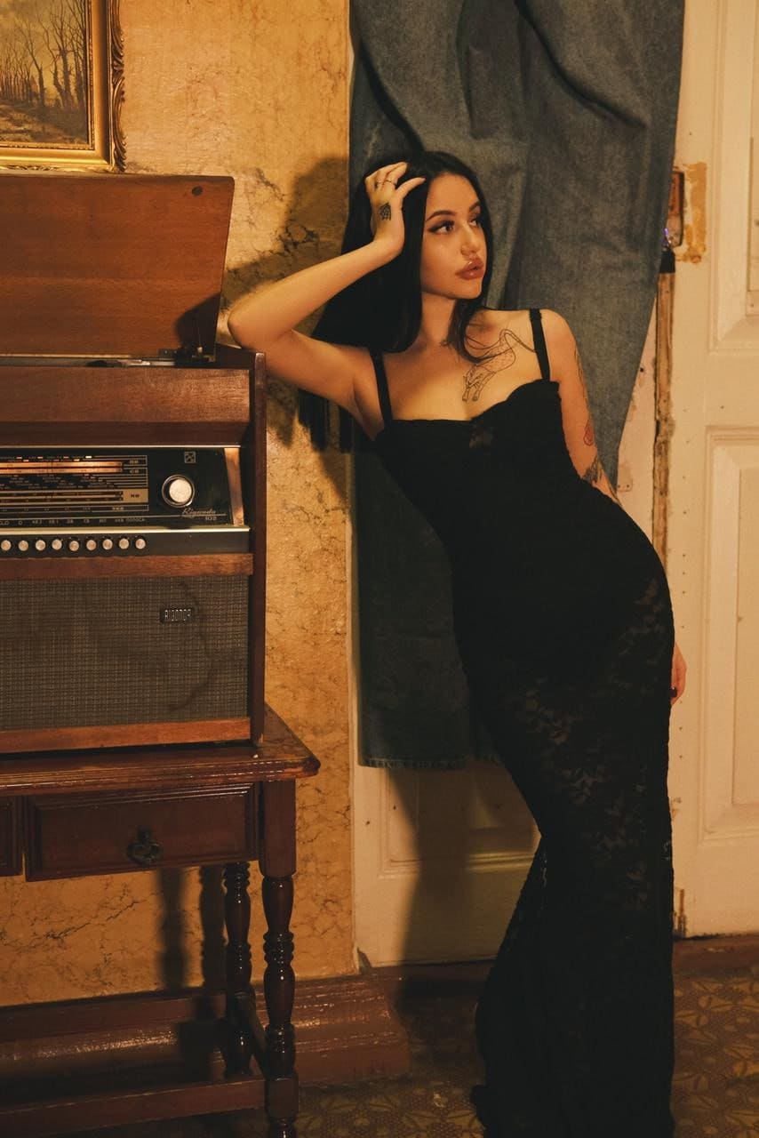 Kate leans against a wall by a wooden cabinet with a vintage radio, wearing a long black lace dress with thin straps and her hand raised to her head.