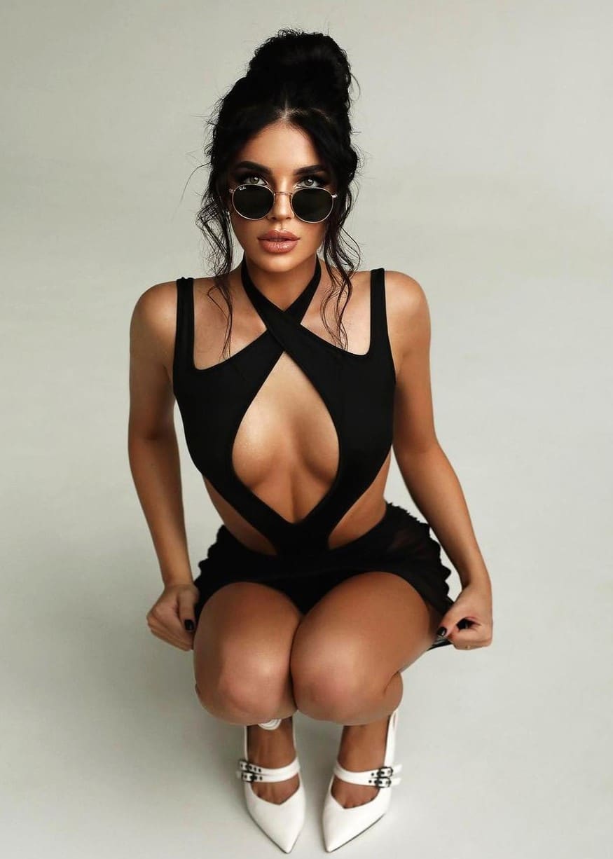 A woman with a high bun crouches in a black cut-out dress and round sunglasses, wearing white pointed-toe shoes on a light background.