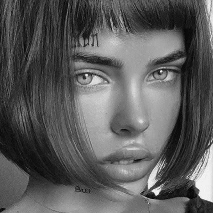 Black-and-white close-up of a person with a bob and bangs, showing forehead tattoos and a dark top against a light background.
