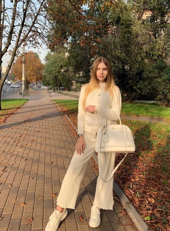 Woman in cream top and wide-leg trousers stands on brick sidewalk in autumn park, holding white handbag, wearing sneakers.