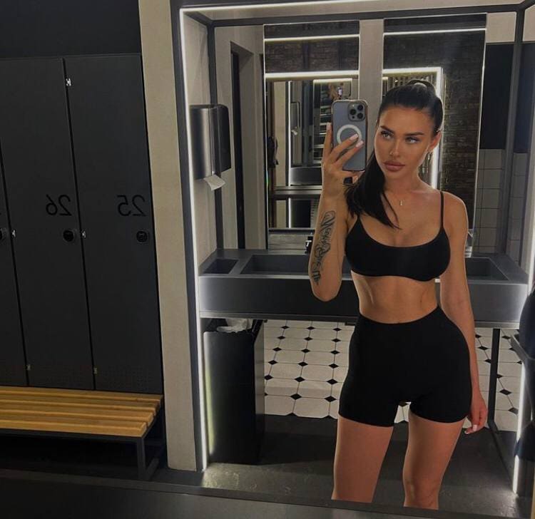 A woman with a high ponytail takes a mirror selfie in a locker room, wearing a black sports bra and shorts.