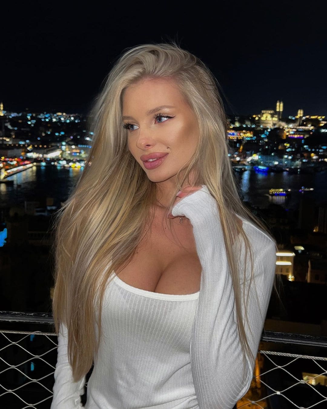 Ella stands on a balcony at night in a white ribbed top and cardigan, with a cityscape and water behind her.