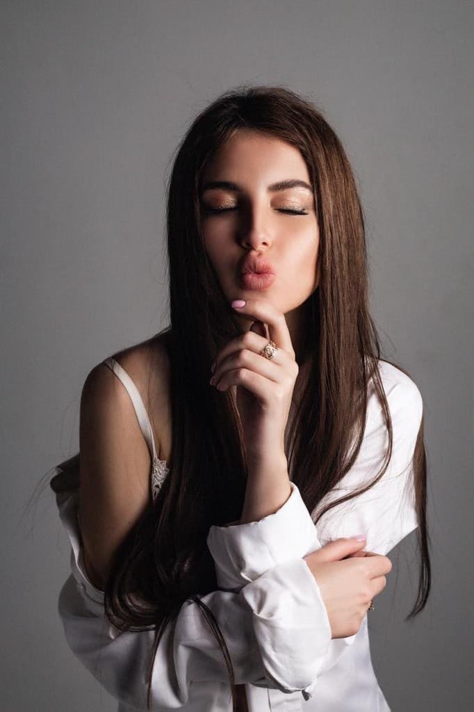A woman in a white shirt slipping off one shoulder poses with eyes closed and lips puckered, finger on chin, against a plain gray background.