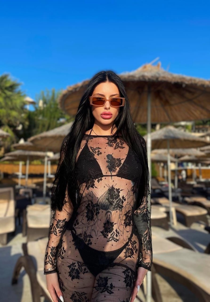 Outdoors by thatched umbrellas, she wears sunglasses and a sheer black lace bodysuit over a black bikini.