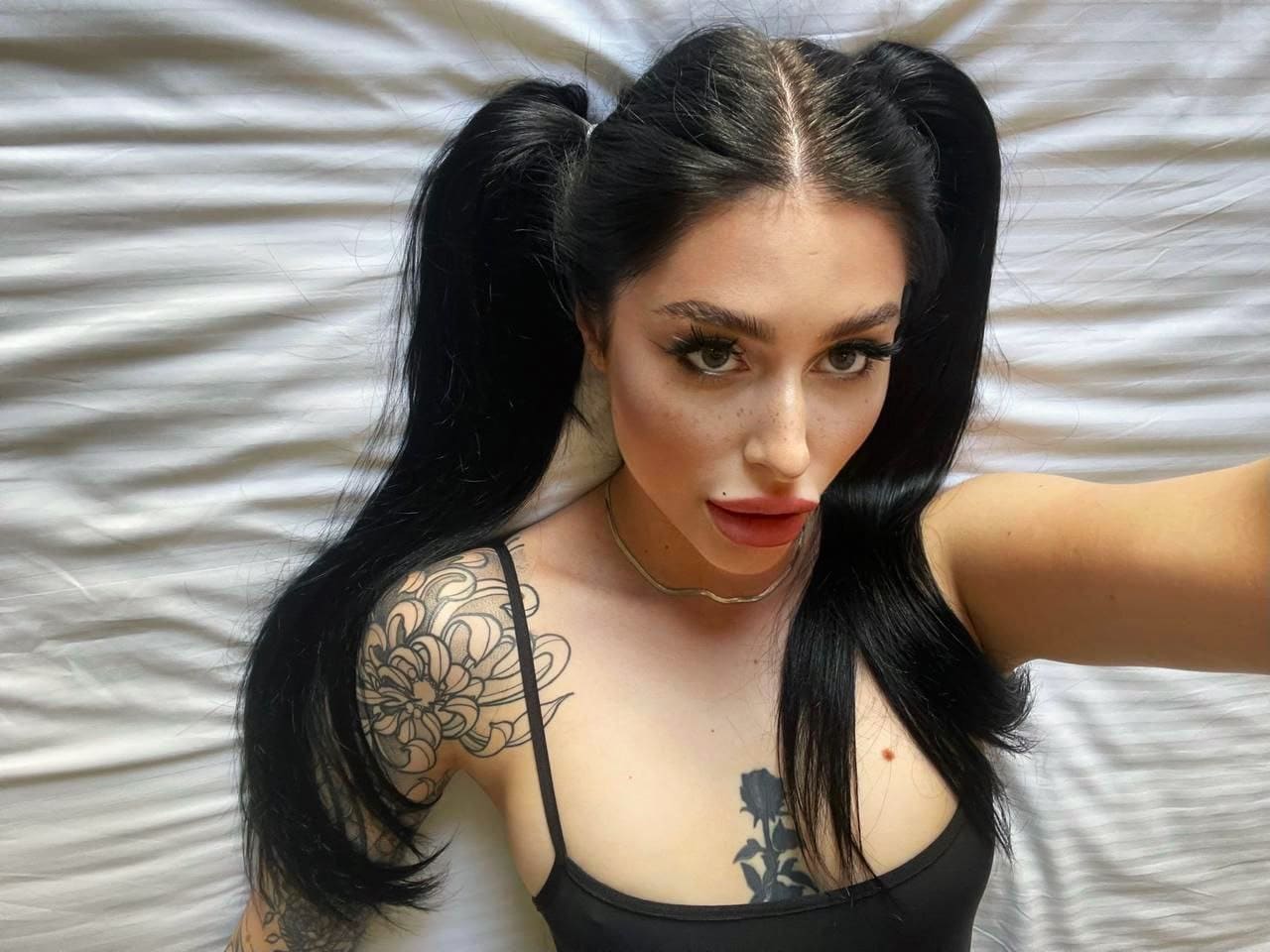 A selfie against a white sheet shows a woman in twin pigtails and a black top, with floral and rose tattoos.
