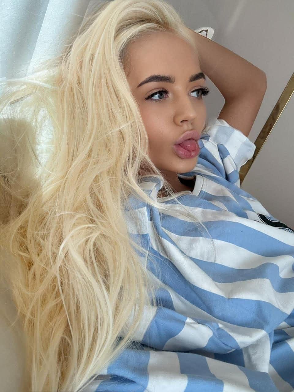 A woman in a blue and white striped t-shirt reclines against a white surface, arm behind her head, sticking her tongue out.