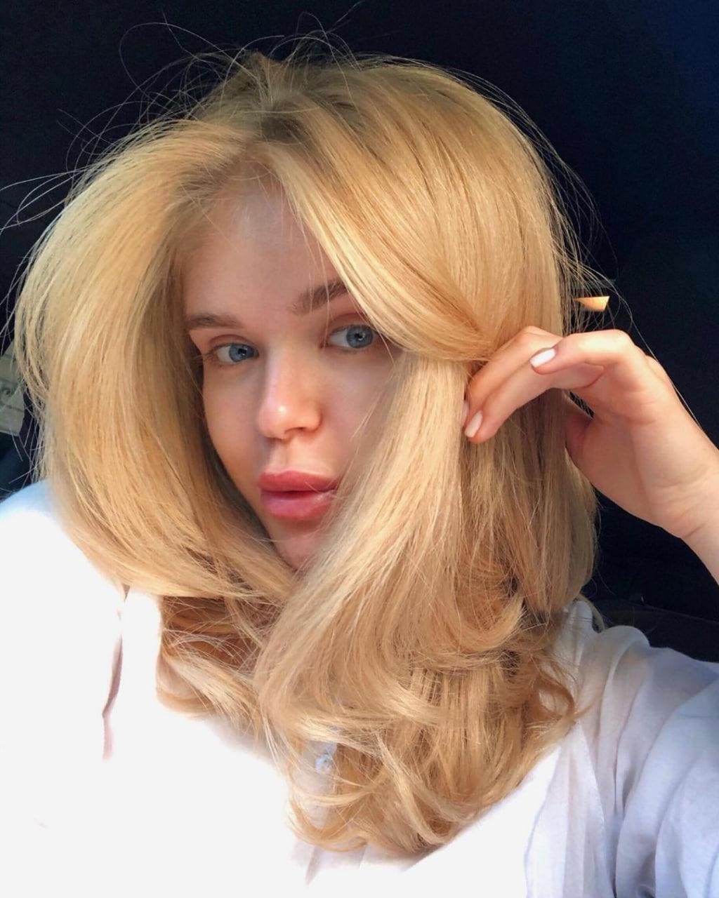 Madelyn with long wavy blonde hair and a white top touches her hair against a dark background.