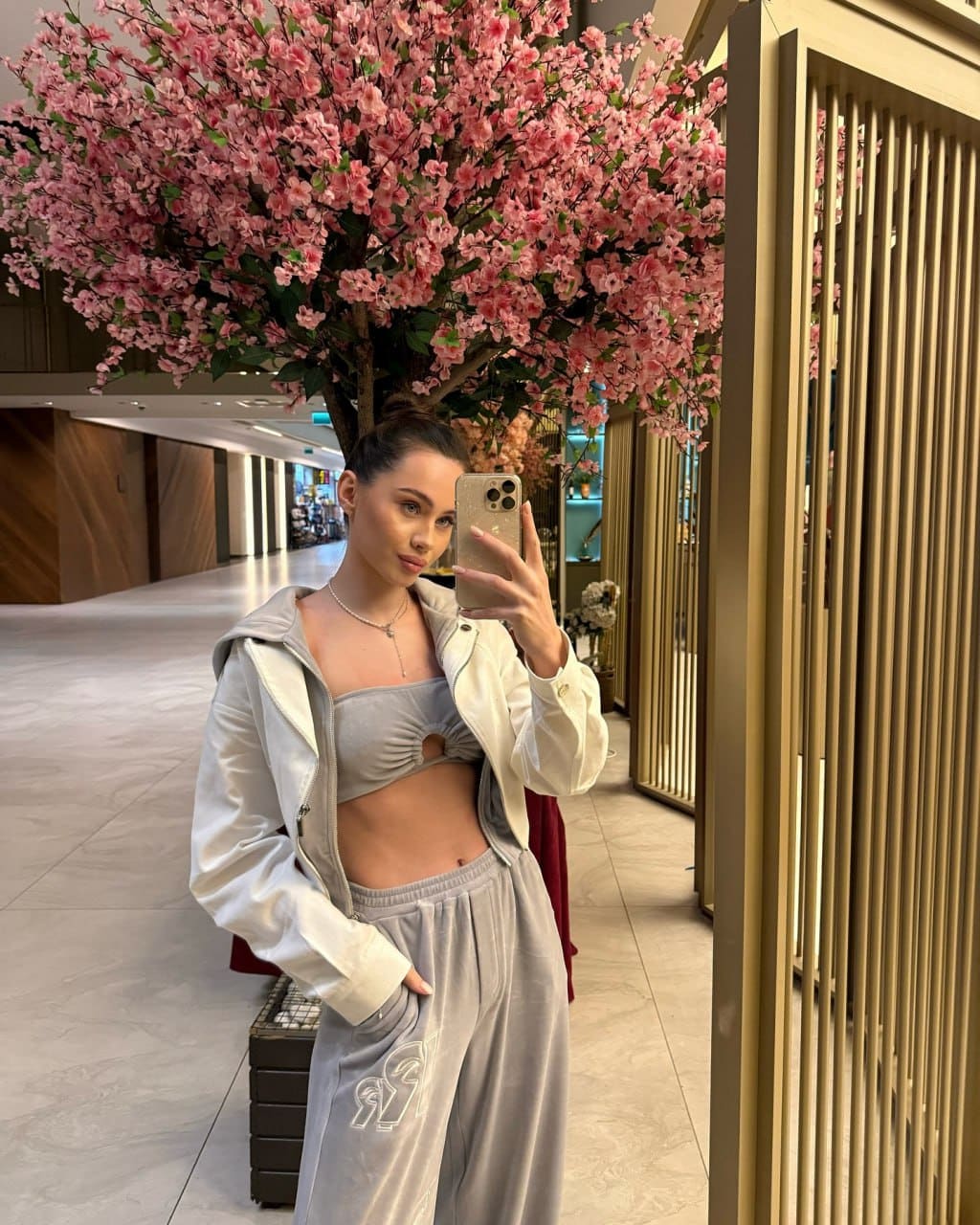 A mirror selfie captures a woman in a grey cropped top, sweatpants, and open white jacket, standing in a corridor before a pink flowering tree.