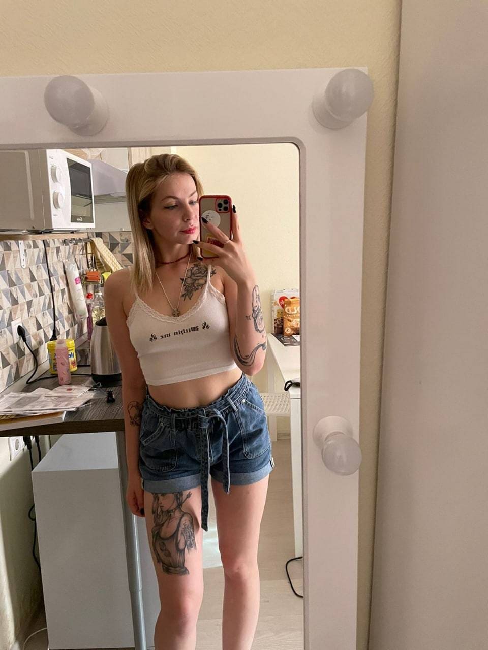 Blonde woman with chest, arm, and thigh tattoos takes a kitchen mirror selfie wearing a white cropped tank and denim shorts, holding a red phone.