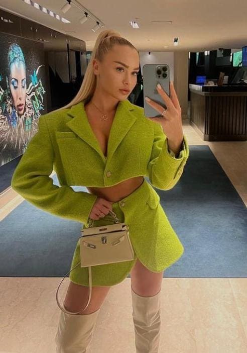 A woman with a high ponytail takes a mirror selfie in a green tweed jacket and mini skirt, beige boots, and handbag, posing with one hand on her hip.