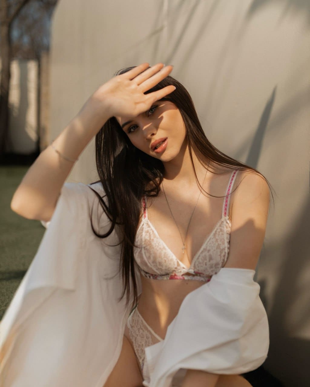 A woman in white lace lingerie with a shirt draped off her shoulders sits outdoors against a light wall, raising a hand to shield her eyes from the sun.