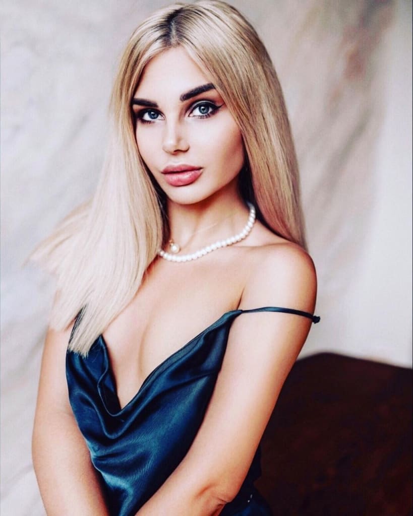Asya sits in a dark teal satin slip dress with a pearl necklace against a neutral backdrop.