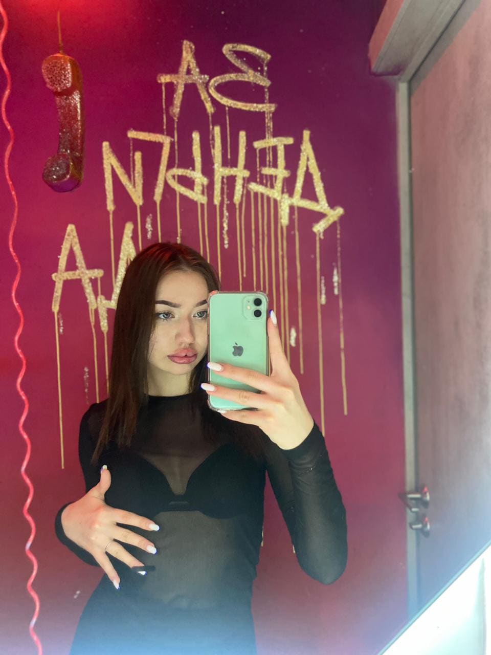 Clara takes a mirror selfie in a black sheer long-sleeve top, standing before a red wall with gold lettering and a hanging phone prop.
