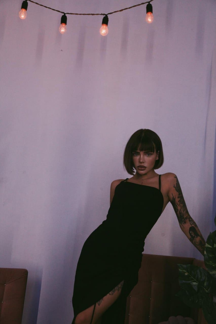 In a dim room, a woman in a black one-shoulder dress leans on a brown sofa, with string lights hanging above against a white backdrop.