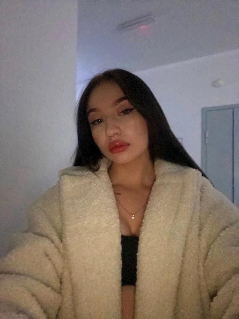 A woman with long dark hair wears a cream fluffy coat over a black top, standing indoors near a closed door with a slight head tilt.