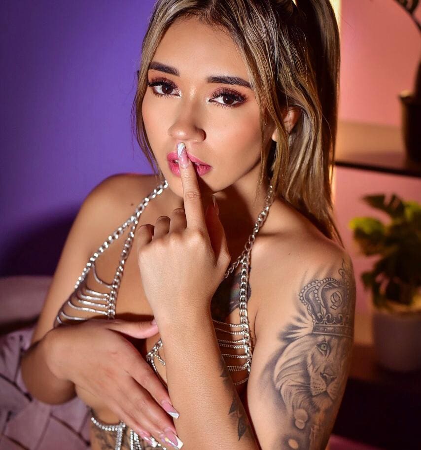 A woman in a silver chain halter top holds a finger to her lips in a softly lit room with purple and pink walls.