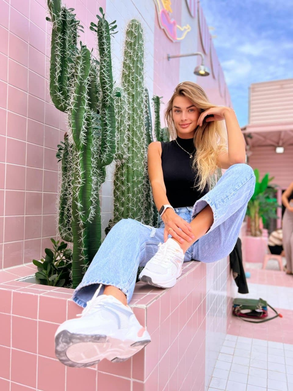 Blonde woman in a black sleeveless top and light blue wide-leg jeans sits on a pink tiled ledge near tall green cacti.