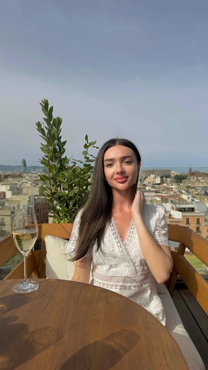 A woman in a white lace dress sits at a rooftop table with white wine, cityscape visible in the background.