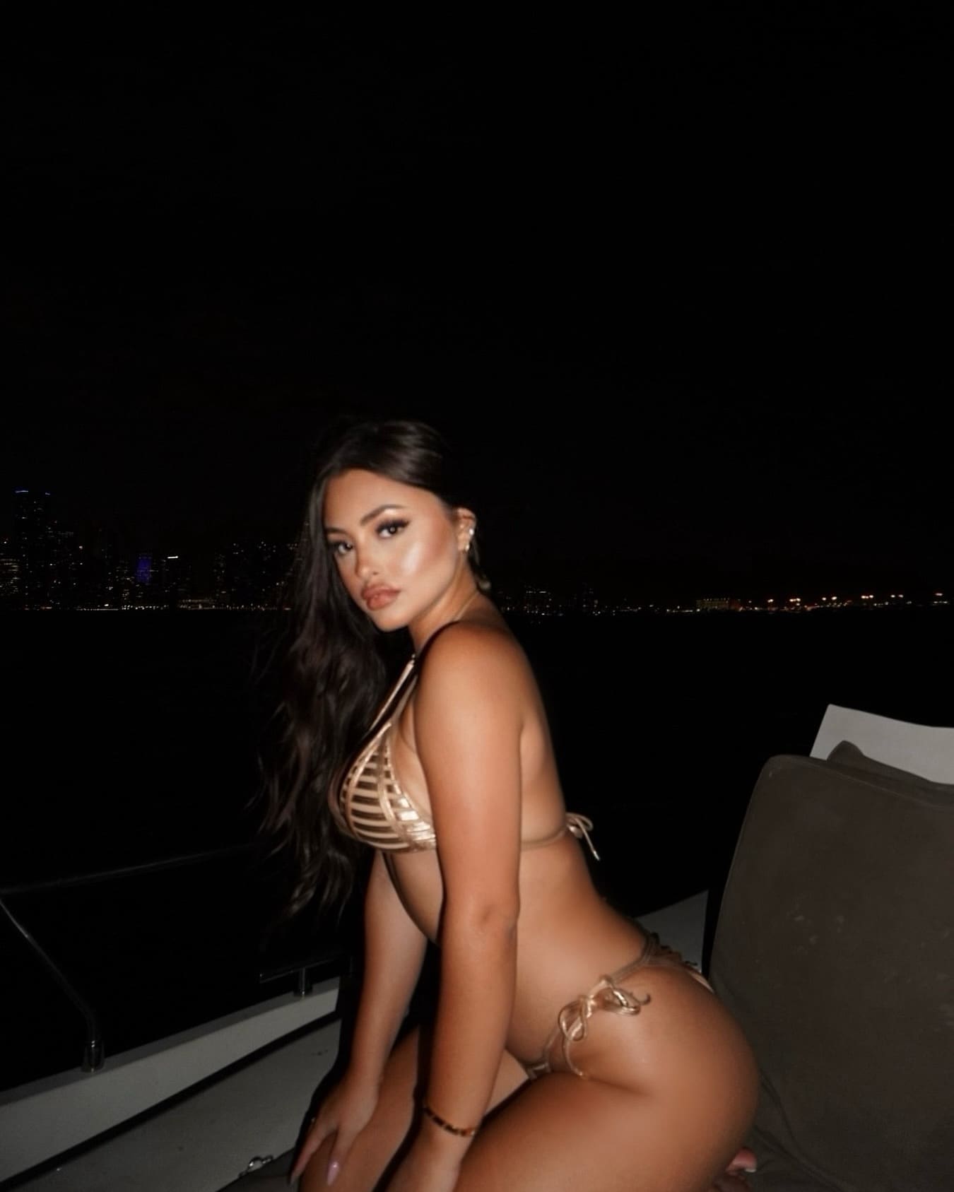 A woman in a gold string bikini sits on a boat at night, with a city skyline visible in the dark background.