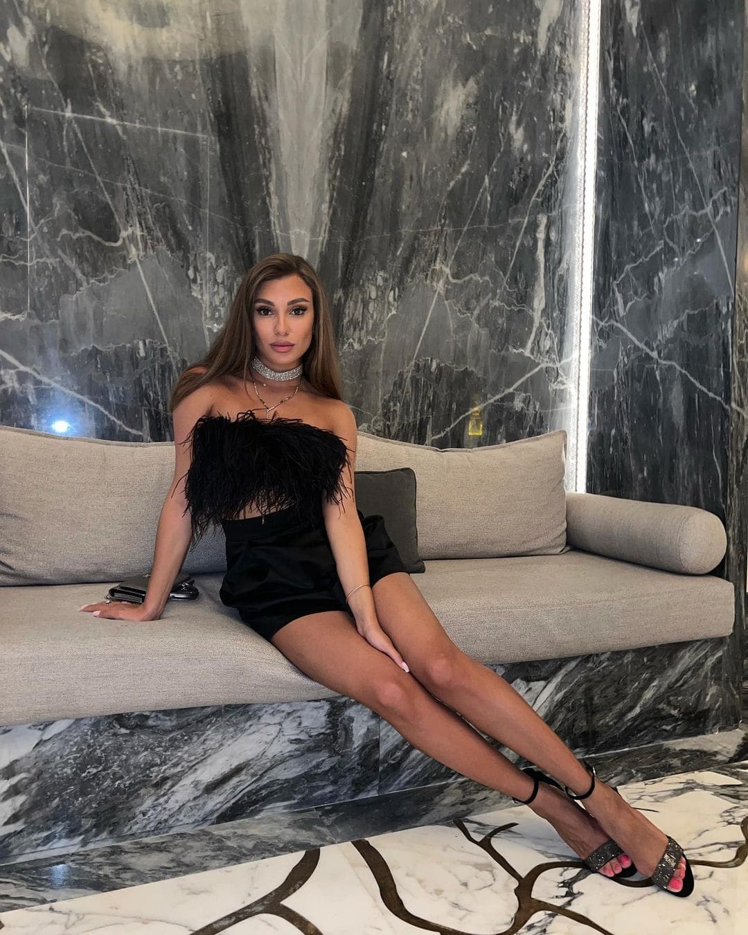 A woman sits on a grey sofa wearing a black feathered top, short skirt, and strappy heels, with pink toenails visible.