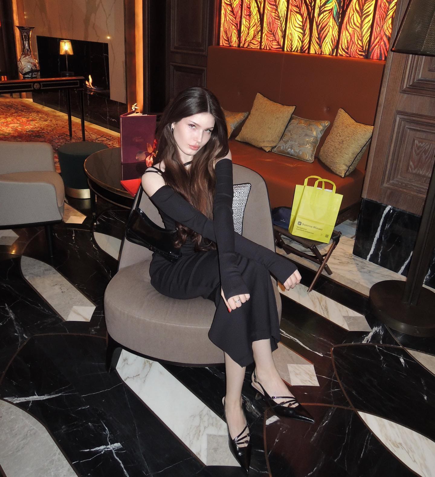 A woman in a black cut-out shoulder dress sits on a beige armchair in a marble lounge, with bags and a gift box nearby.
