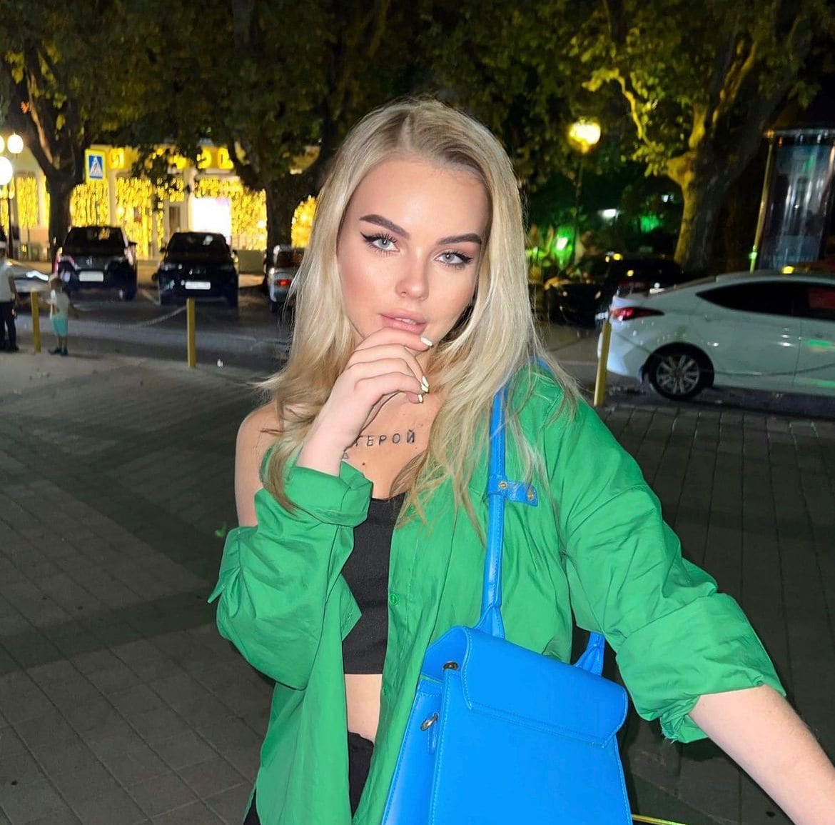 A blonde woman stands on a city sidewalk at night in an open green shirt and black crop top, hand to chin.