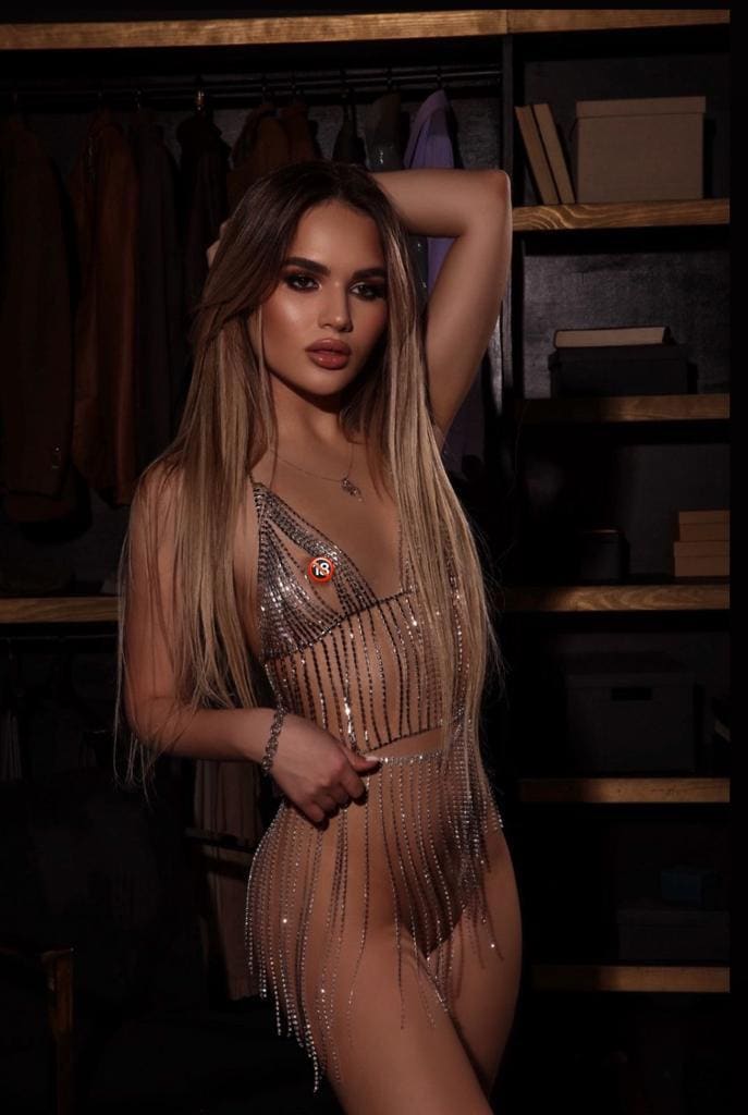 Nita stands in a dim room with shelves, wearing a sheer beaded fringe bodysuit and raising one arm behind her head.