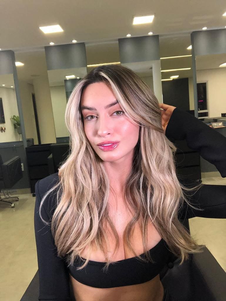 A blonde woman with dark roots wears a black long-sleeved crop top, posing with a hand in her hair in a salon setting.