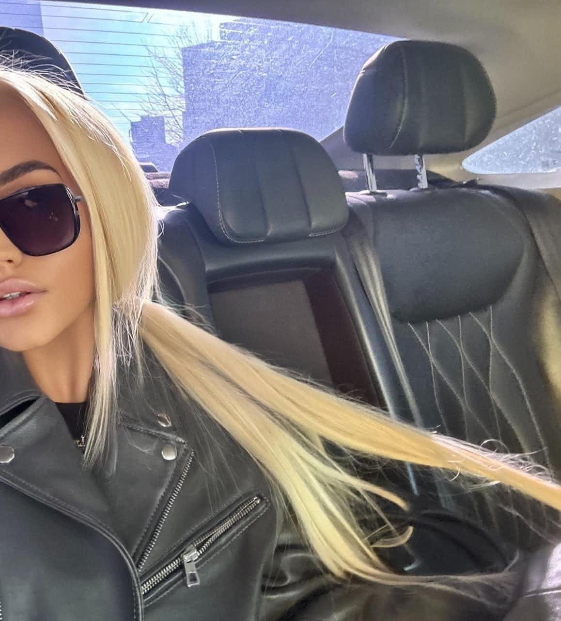 A woman in dark sunglasses and a black leather biker jacket with silver zippers sits in a car's back seat.
