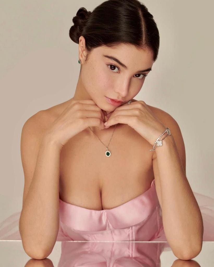 A woman in a strapless pink dress rests her hands under her chin while seated at a reflective table.
