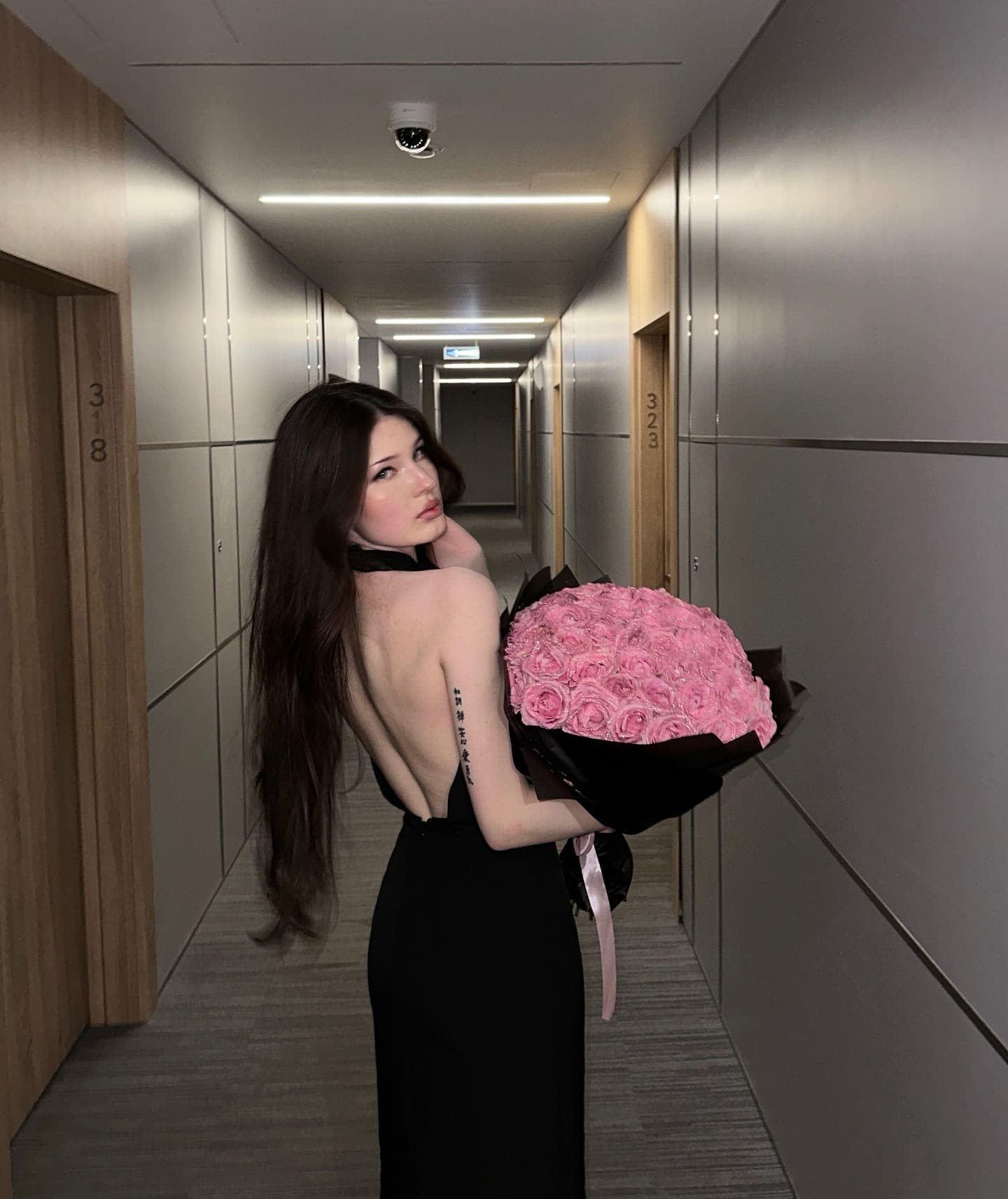 A woman in a black backless dress holds a large bouquet of pink roses while standing in a hotel corridor.