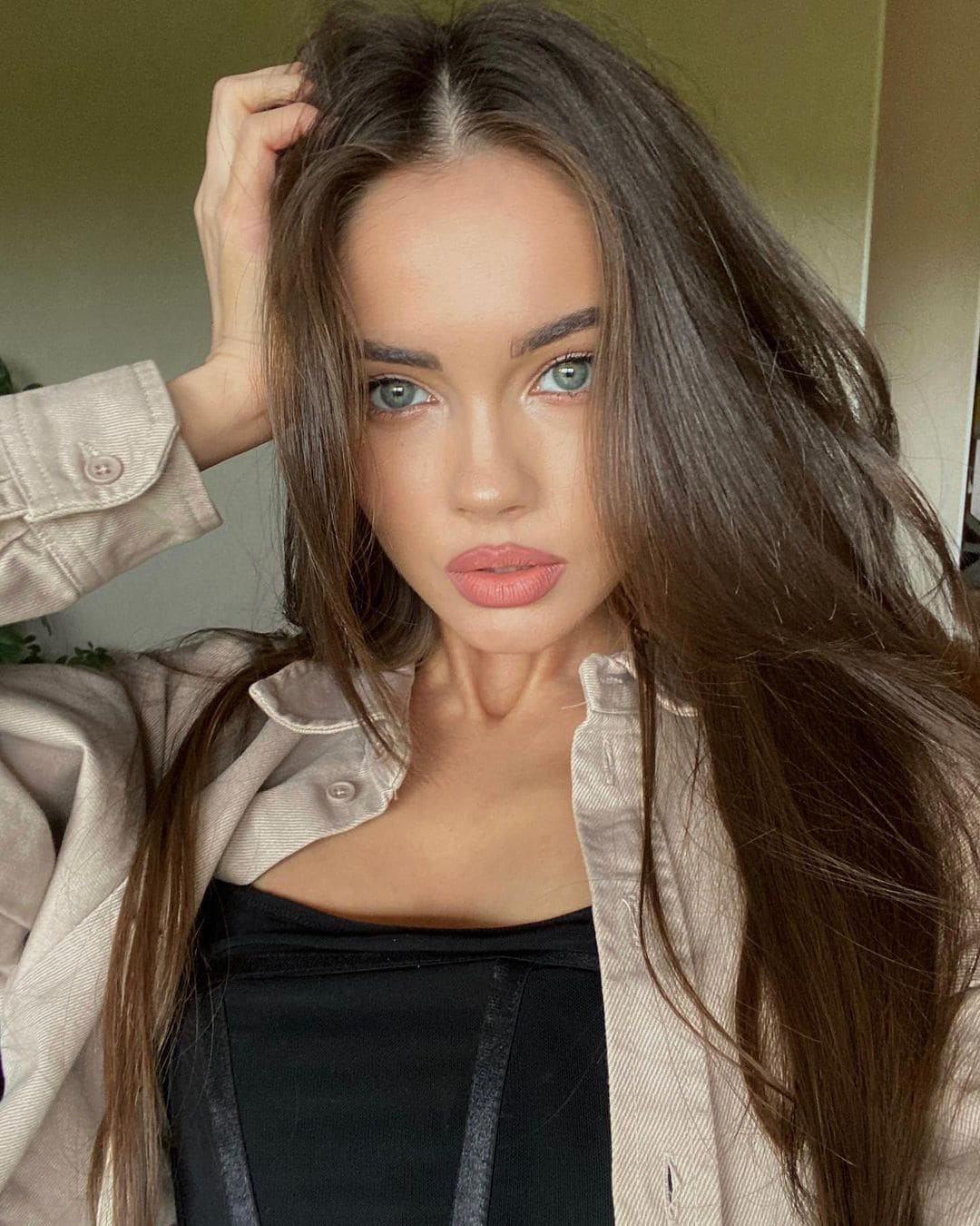 A woman with blue eyes wears a black top under an open light button-up shirt, raising a hand to her hair while posing indoors.