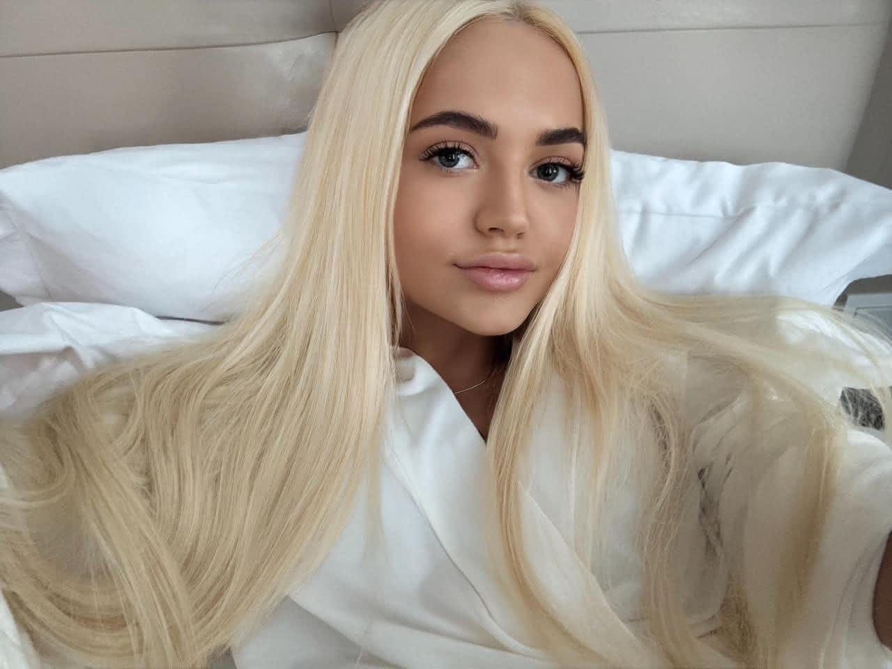 Sveta reclines against white pillows on a bed, wearing a white collared shirt or robe.