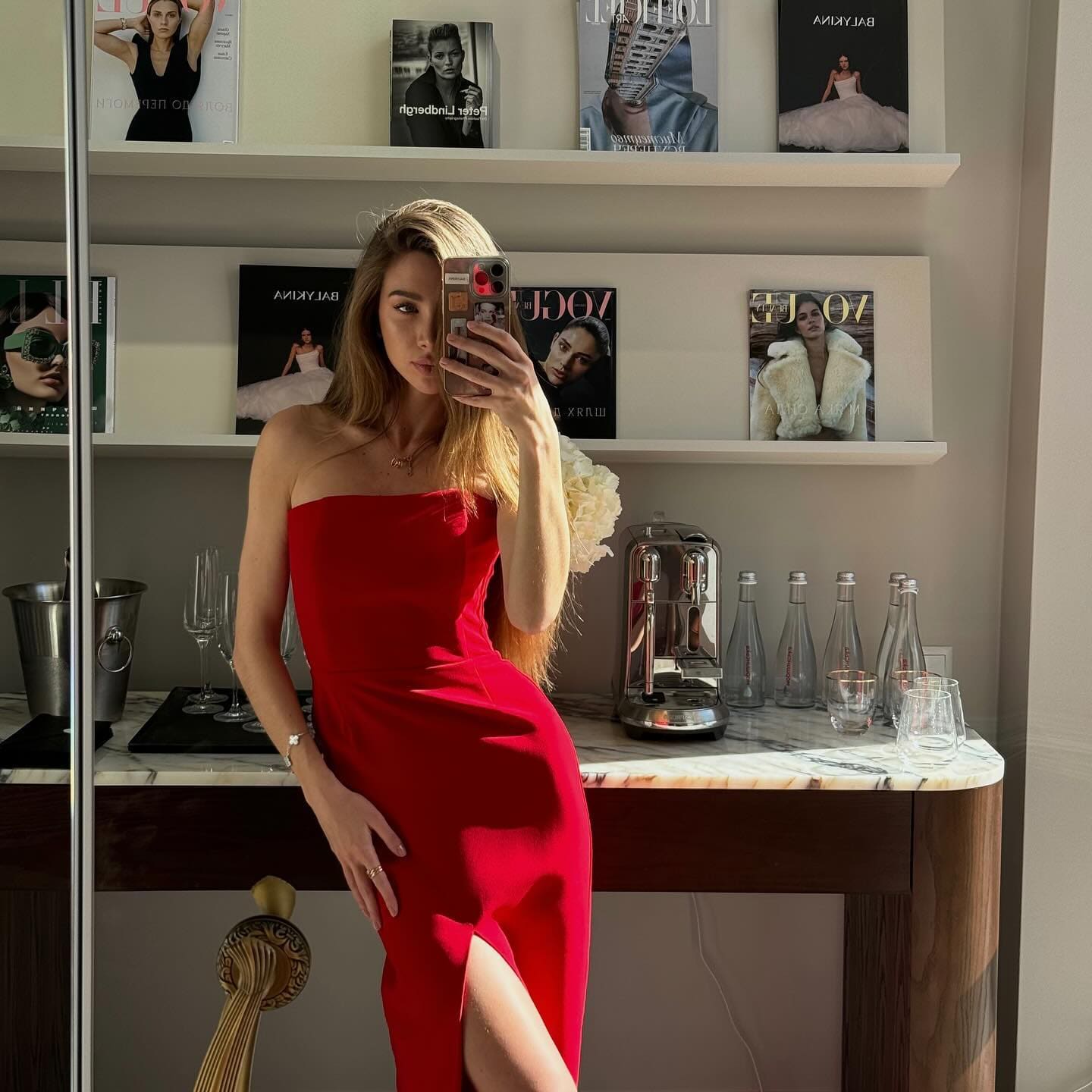 A blonde woman takes a mirror selfie in a strapless red dress with a slit, in a room with magazine shelves and a coffee machine.